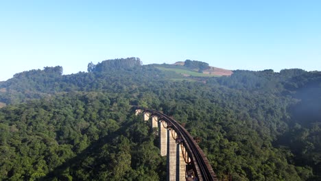 Vista-Aérea-De-La-Vía-Del-Puente-Ferroviario-Alto-A-Través-De-Un-Valle-Hacia-Una-Colina-Con-Bosque-Nativo-Bajo-La-Niebla-De-La-Mañana