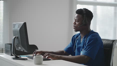 Afro-american-male-doctor-or-nurse-with-headset-and-computer-working-at-hospital-.young-professional-therapist-doctor-consulting-customer-client-using-remote-communication-speaking-on-webcam