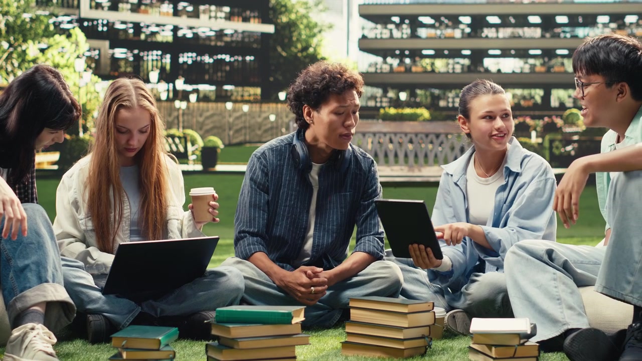 Diverse students checking notes on tablet and laptop with stack of books