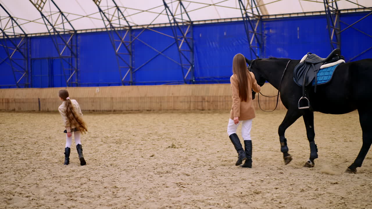 Slim brunette lady walks leading the black horse by the large manege. Young girl comes to a woman taking her by the hand.