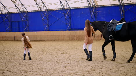 slim brunette lady walks leading the black horse by the large manege. young girl comes to a woman taking her by the hand.