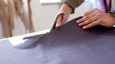 hands of biracial female fashion designer cutting cloth with fabric shears on table, slow motion