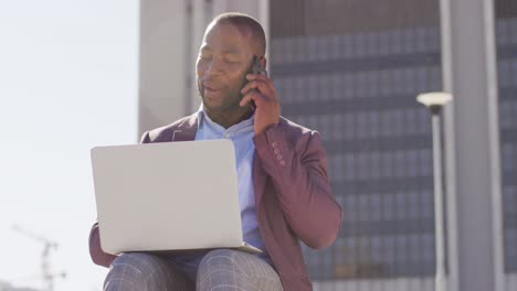 Hombre-Afroamericano-En-La-Ciudad-Sentado-En-Las-Escaleras,-Usando-Un-Teléfono-Inteligente-Y-Una-Computadora-Portátil