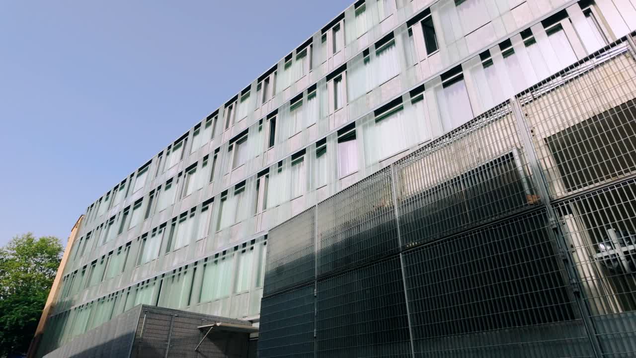 Fachada de edifício moderno com painéis de vidro fosco e grades de metal robustas, vista de um ângulo inferior contra um céu azul claro. Reflete a arquitetura urbana contemporânea.