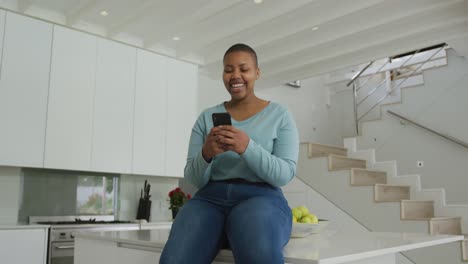 Happy-african-american-plus-size-woman-sitting-on-countertop-in-kitchen,-using-smartphone