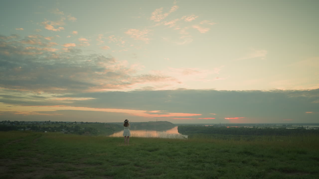 색 드레스와 모자를 입은 여자가 열린 풀에서 혼자 서서 조용한 호수와 멀리 떨어진 마을을 바라보고 있습니다.