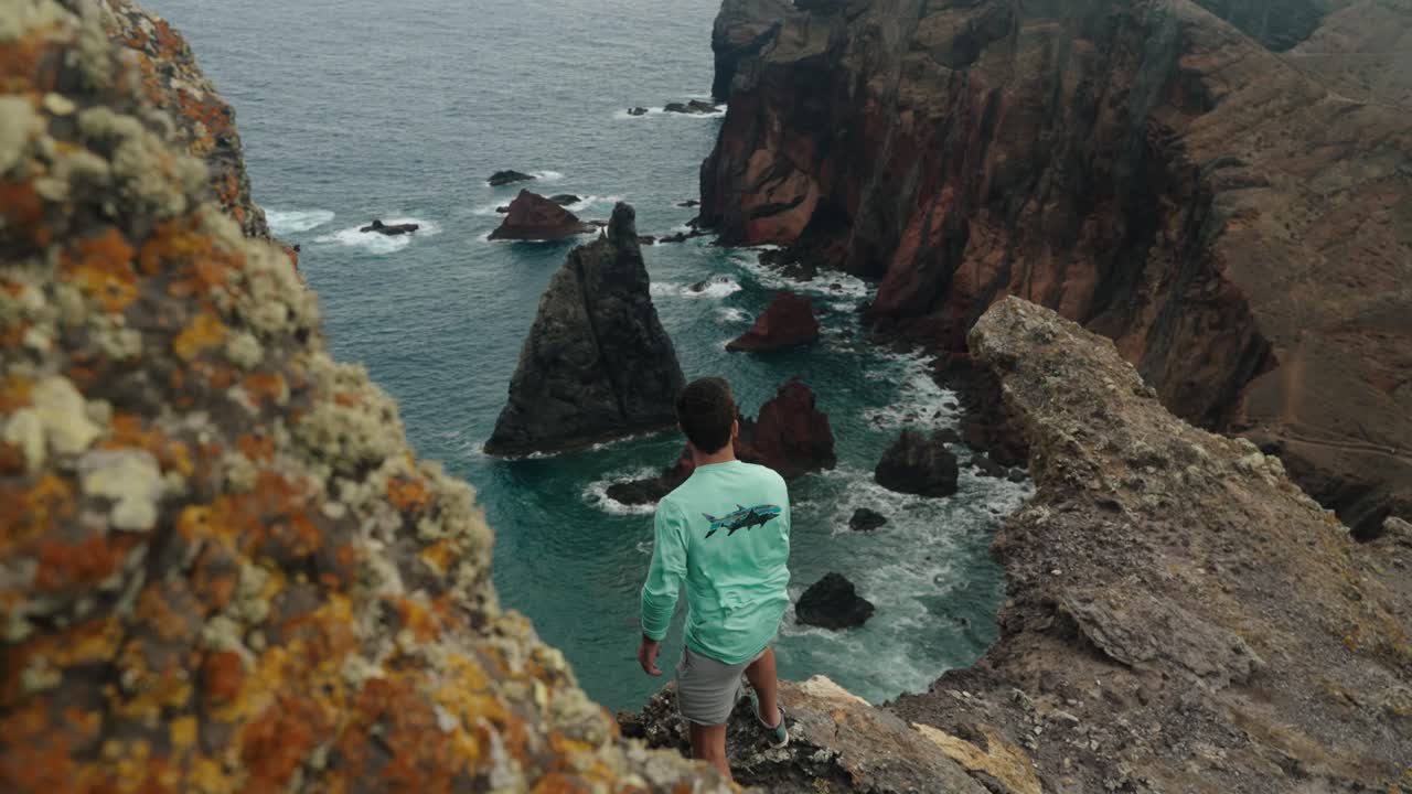 A man stands on the edge of a rocky cliff, overlooking the jagged volcanic coast and turquoise sea below.