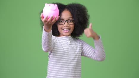 young cute african girl with afro hair holding piggy bank and giving thumbs up