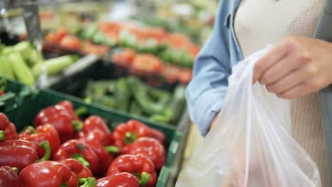 Handheld-footage-of-a-girl's-hands-selecting-fresh-red-sweet-peppers-from-the-box-in-vegetable-department.-Grocery-store.-Girl-puts-bell-peppers-to-the-cellophane-bag