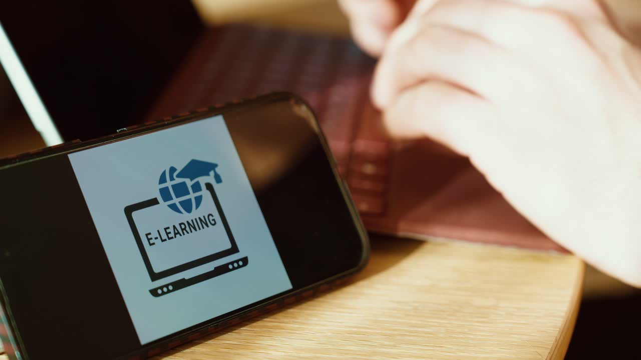 Close-up of a smartphone displaying an e-learning interface while hands type on a laptop at a desk, showing modern online training and remote education. Ideal for corporate learning, upskilling, compl