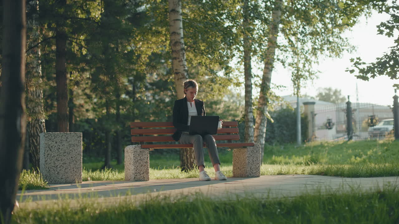 Office assistant seated outdoor in lush park working on system while sipping fruit juice with sunlight filtering through trees, iron fence in background and parked car