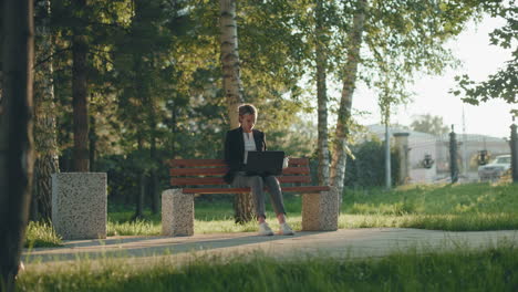office assistant seated outdoor in lush park working on system while sipping fruit juice with sunlight filtering through trees, iron fence in background and parked car