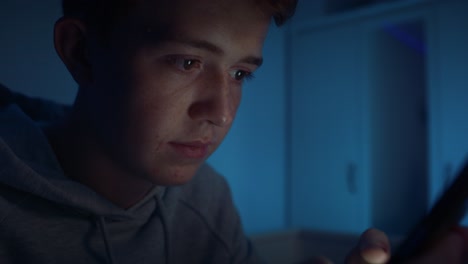 un adolescente caucásico usando un teléfono móvil mientras está sentado por la noche en su habitación.