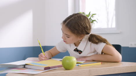 Pretty-little-girl-in-uniform-sitting-at-desk-and-writing-in-notebook-during-English-class-at-school