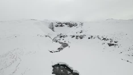 Toma-Aérea-De-Un-Hombre-Caminando-Por-Montañas-Nevadas-Hacia-La-Cascada-Svöðufoss-En-La-Península-De-Snæfellsnes,-Islandia