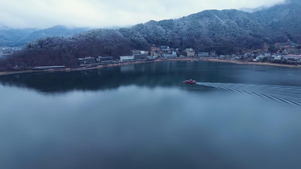 Phong cảnh trên không từ drone về một chiếc thuyền địa phương đang di chuyển qua vịnh hồ Kawaguchi với những cây rừng phủ tuyết trên mũi núi và các tòa nhà thị trấn nông thôn ở phía sau, Yamanashi, Nhật Bản, du lịch châu Á