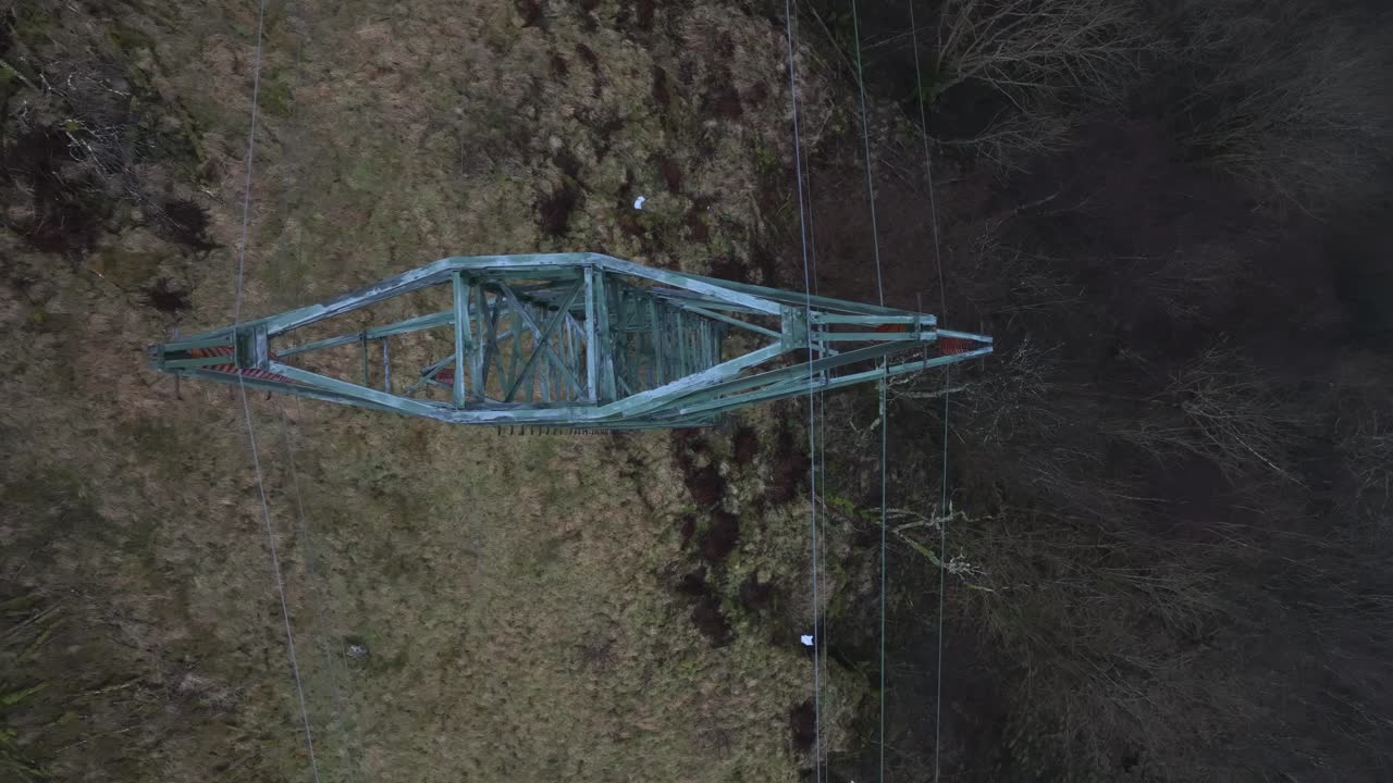 Aging high voltage pylon with power cables shows signs of corrosion. Drone moves slowly sideways in a birdseye aerial view, capturing infrastructure in misty, rainy weather