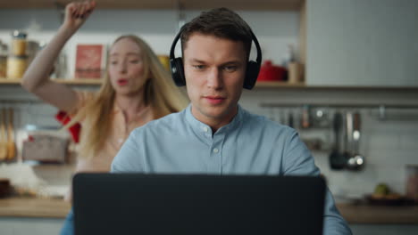 manager consulting online web camera in headphones at kitchen while wife dancing