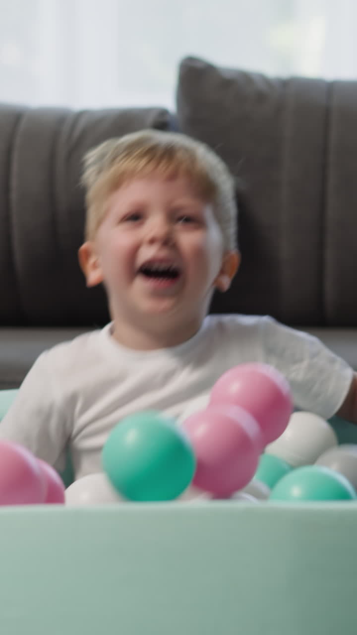 Laughing little children jump into soft pool with plastic colorful balls in living room slow motion. Active kids enjoy game at home closeup