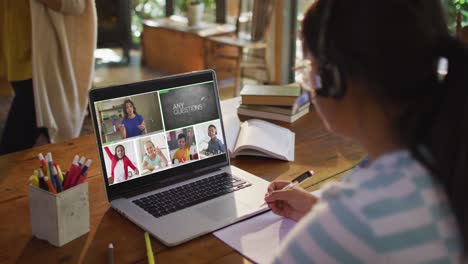 Schoolgirl-using-laptop-for-online-lesson-at-home,-with-diverse-teacher-and-class-on-screen