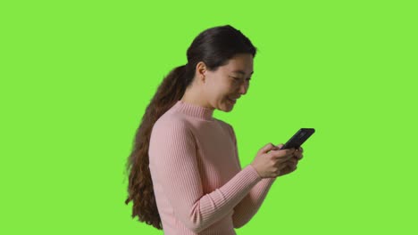 studio shot of woman smiling and laughing at message or content on mobile phone walking in front of green screen