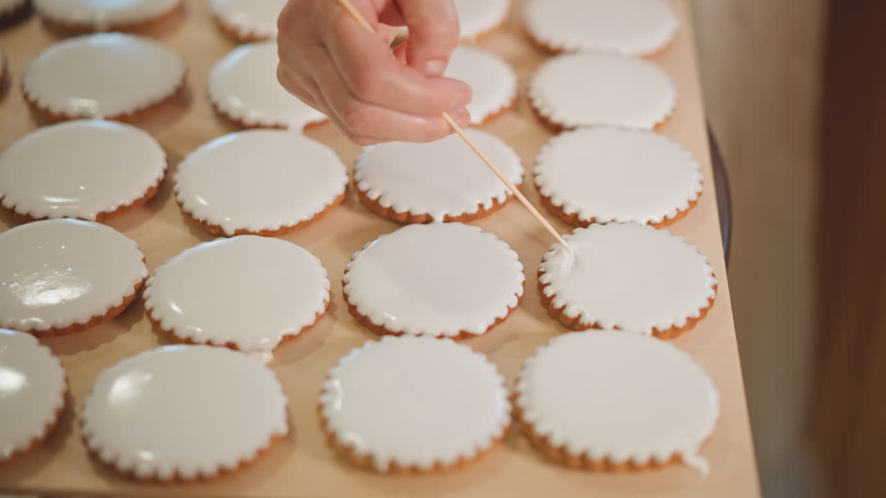 Iced sugar cookies on wooden tray, pastry chef decorating dozens with toothpick. Close view of neat round cookies with smooth white glaze as hand dots and adjusts surface, warm ambient bakery light