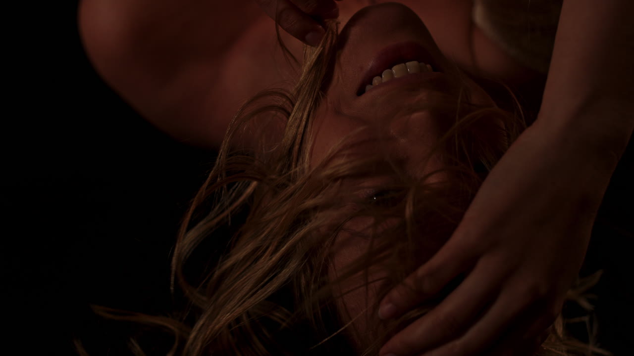 Beautiful portrait of a happy, young woman sensually playing with her blonde hair in slow motion in a dark room