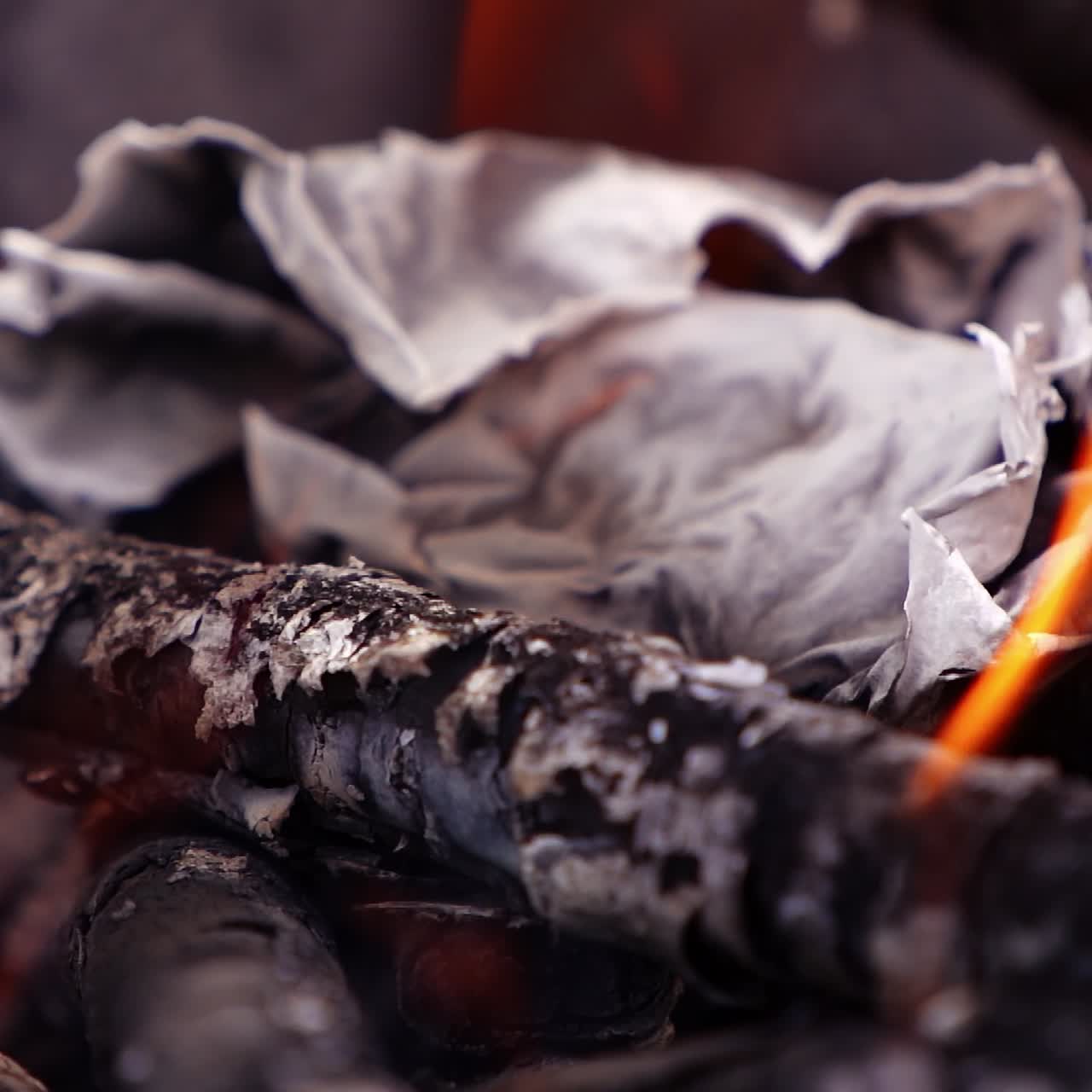 Fire tongues over the smouldering logs. Paper twisting in fire. Embers of burnt-out wooden logs in slight flame. Close-up. Slow motion.