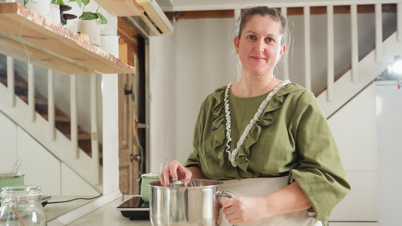Lady in green gown stands in cozy kitchen, dipping electric mixer into bowl and pulling it out with thick white dough as she smiles gently and looks directly into camera in warm natural light setting