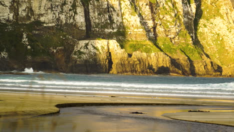 Statische-Aufnahme-Von-Wellen,-Die-Auf-Den-Strand-Von-Purakaunui-Krachen,-Mit-Einem-Riesigen-Berghang-Dahinter