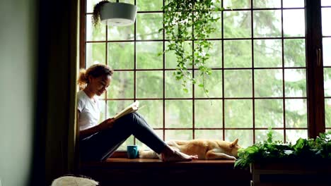 attractive girl in casual clothes is reading book sitting on window sill barefoot near adorable pet dog lying near her owner. home, hobby and animals concept.