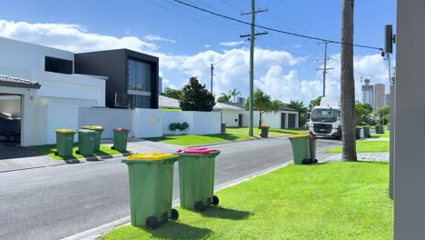 a modern garbage truck uses a robotic arm to empty residential trash and recycling bins along a sunny suburban street, captured in bright daylight with smooth camera movement