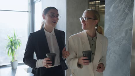 two confident businesswomen in black and white uniforms and glasses come out of the elevator while starting their working day drink coffee and chat while walking around a modern office