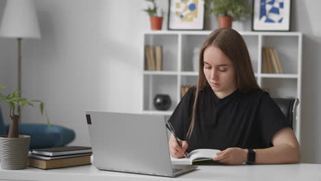 Die-Junge-Frau-Liest-Informationen-Auf-Dem-Laptop-Bildschirm-Und-Schreibt-In-Ein-Notizbuch,-Um-An-Einem-Online-Fernkurs-Für-Die-Berufliche-Weiterbildung-Zu-Hause-Zu-Lernen