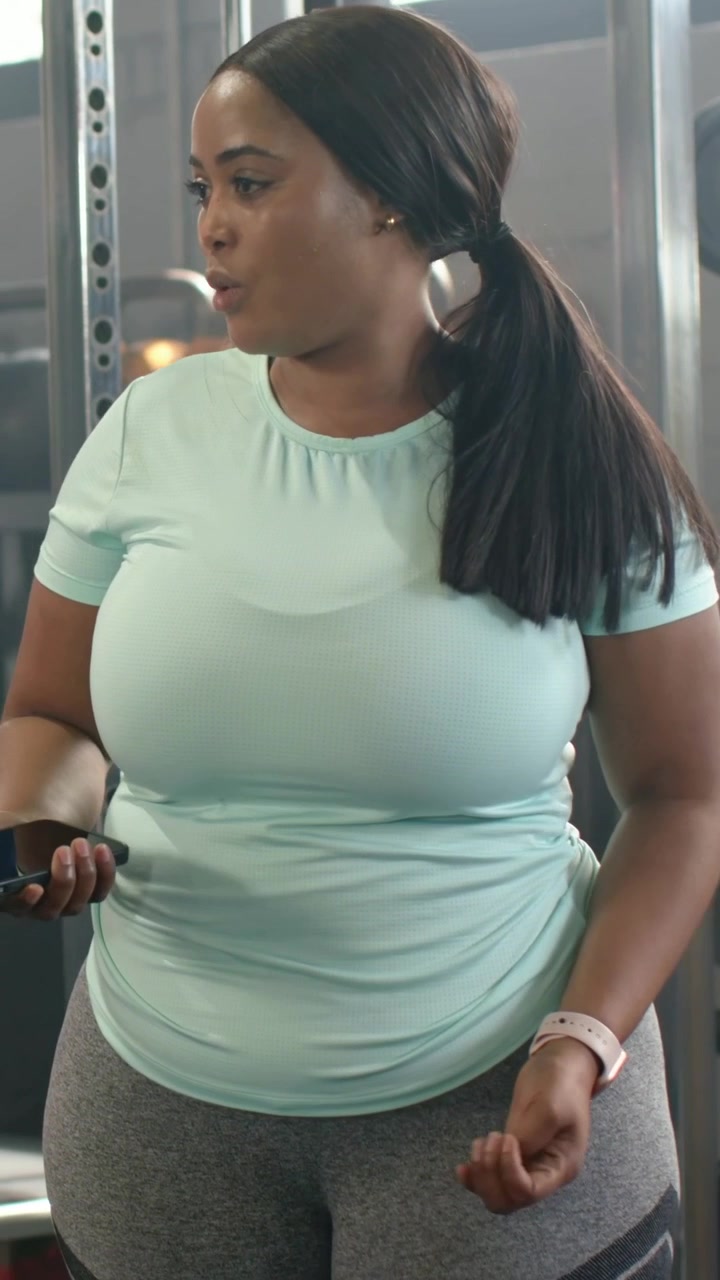 Vídeo vertical: Mulher de blusa menta segurando o telefone e conversando, verificando o treino no rack da academia