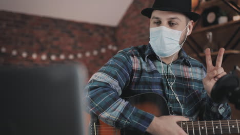 joven músico masculino con mascarilla médica grabando y transmitiendo un video tutorial, tocando la guitarra en casa durante el encierro debido a la pandemia de covid 19