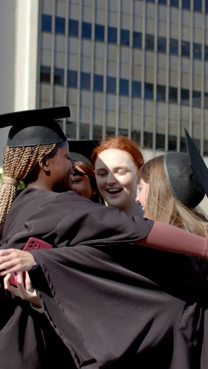 Vertical video: Lifting diploma, four in gowns celebrating and hugging on campus, caps visible