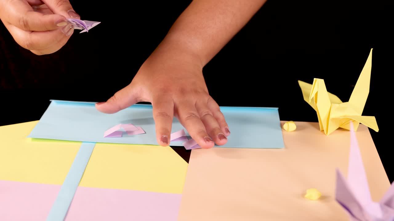Hands remove small paper craft from pastel table, surrounded by origami, under bright studio lighting