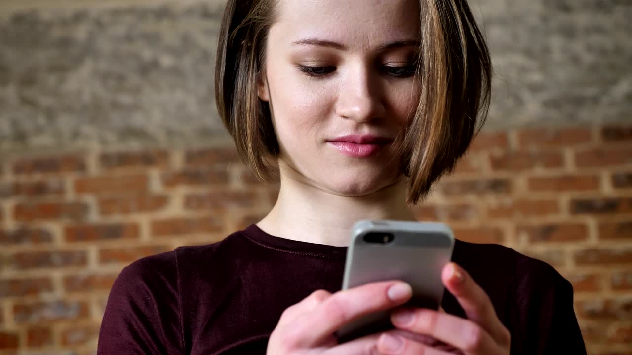 Young beautiful girl is reading message on her smartphone, laughing, communication concept, brick background