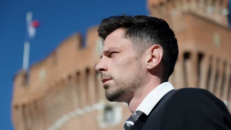 young man in formal wear gazes ahead with confidence near historic brick tower under blue sky