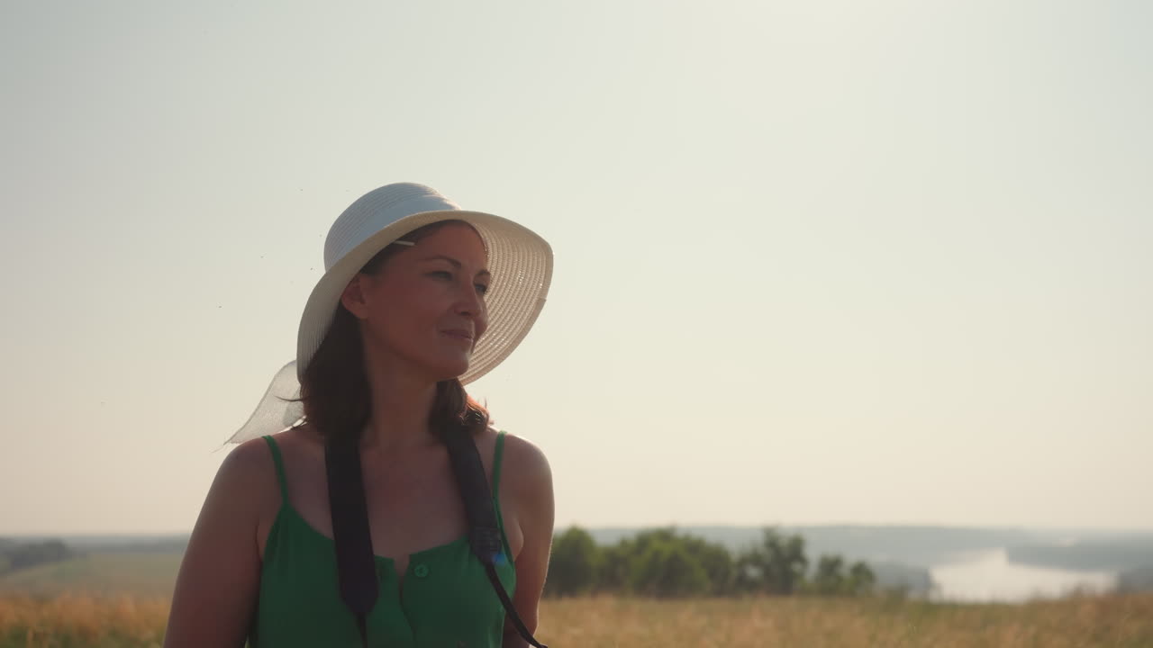 white woman in sunhat and green dress walks through dry farmland holding DSLR camera gazes at blurred distant landscape under golden light capturing rural environment photography