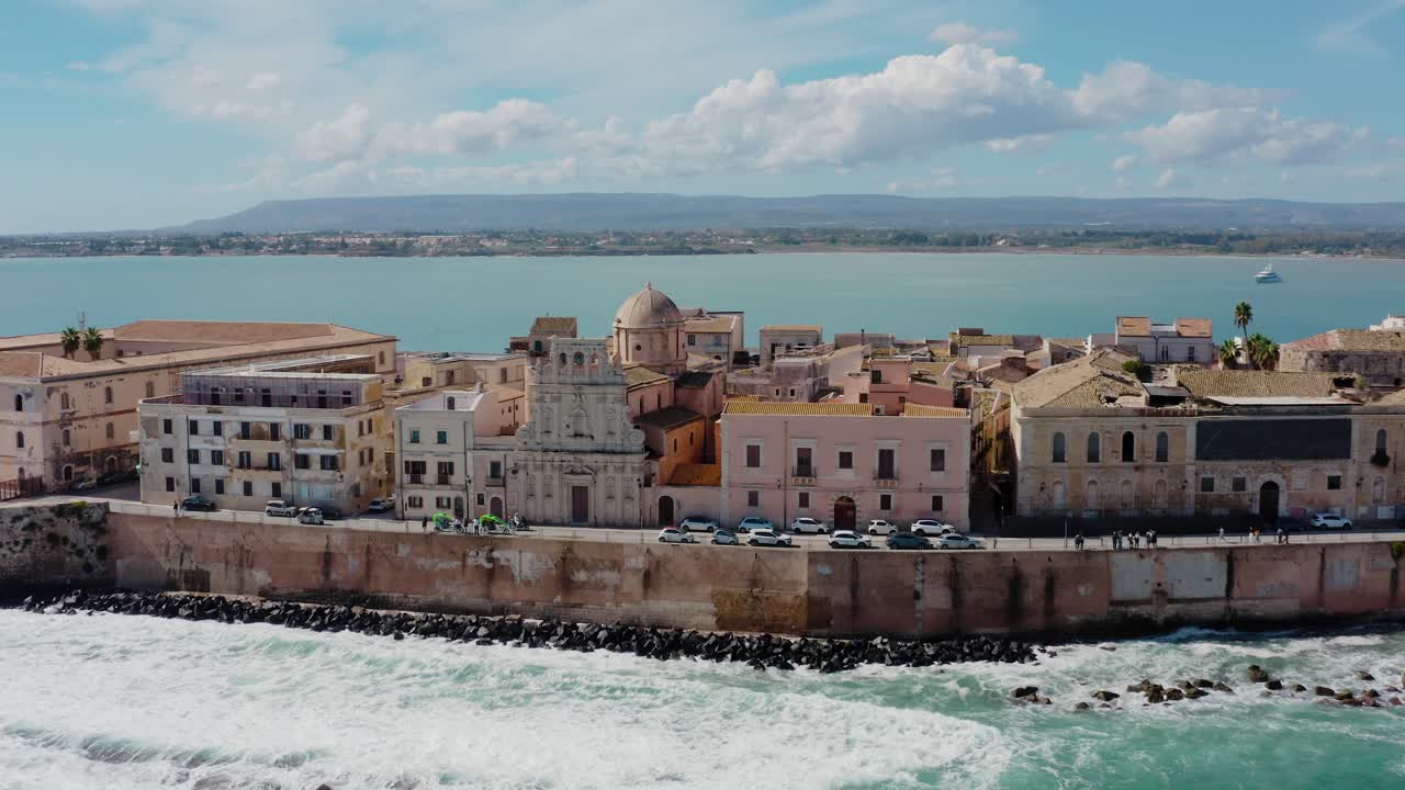 Aerial drone shot of historic buildings on the island of Ortigia with waves crashing on the rocks. Medieval town in Sicily. Old town of Syracuse.