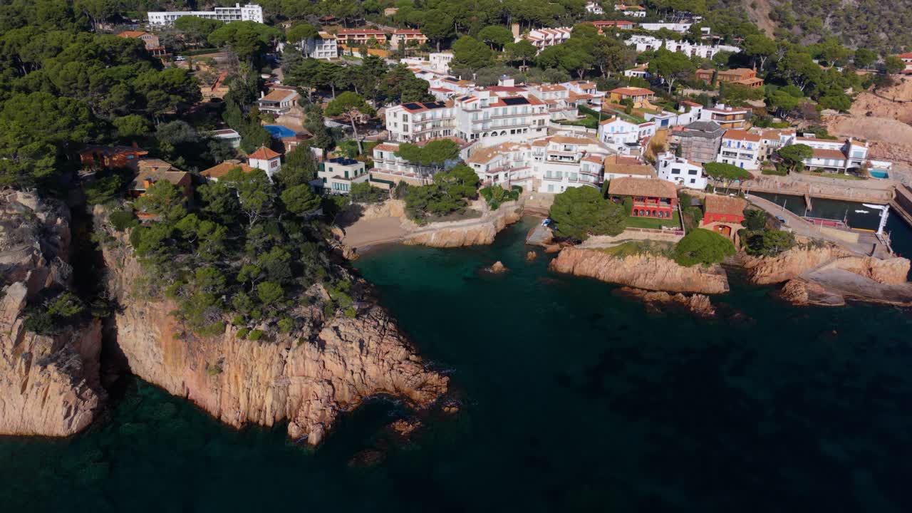 Drone footage ascending from the secluded cove of platja d'en malaret, revealing the picturesque coastal village of fornells with its traditional white houses nestled along the rocky coast
