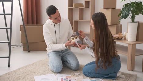 young couple in a new house sitting on the carpet and choosing colours for decoration wth a house model