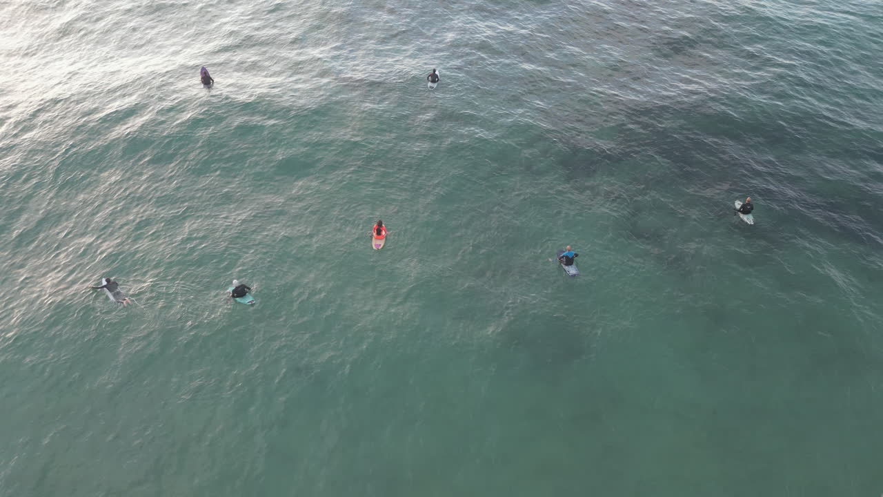 toma aérea de drones inclinándose hacia arriba para revelar un grupo de surfistas esperando atrapar la próxima ola perfecta mientras el sol se pone en la distancia sobre el mar cantábrico, suances, españa