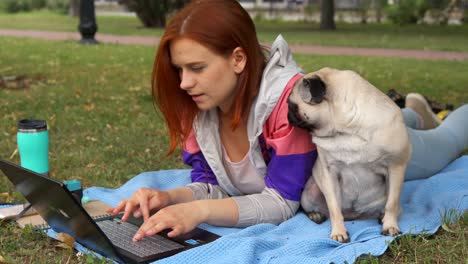 girl laying and typing on laptop on a lawn with her pug around