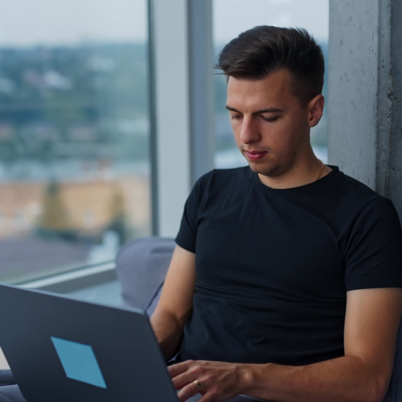 Dark-haired young man focused on his work at laptop. Male sitting near the panoramic window in a comfortable chair. Blurred backdrop