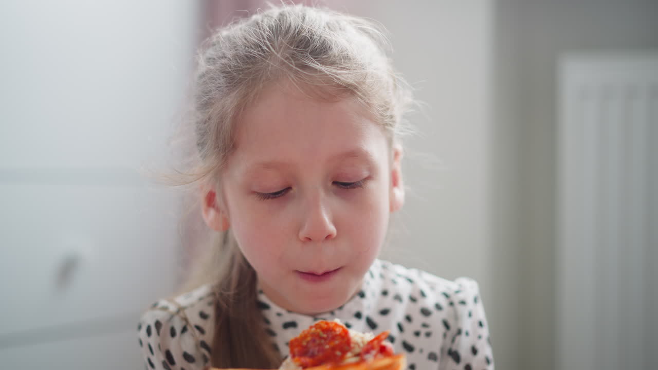 Cute girl holding grilled pepperoni pizza slice looks attentively at someone offscreen while seated in brightly lit cozy indoor space, enjoying moment of shared mealtime with warm daylight ambiance