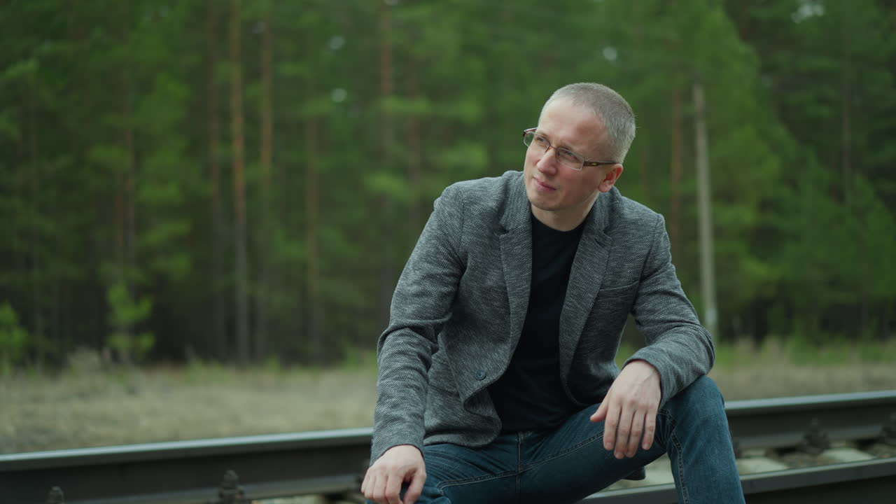 una vista de cerca de un hombre con una chaqueta gris ajustando sus gafas sentado en una vía ferroviaria, mirando pensativo, el fondo presenta árboles verdes y vías ferroviarias borrosas