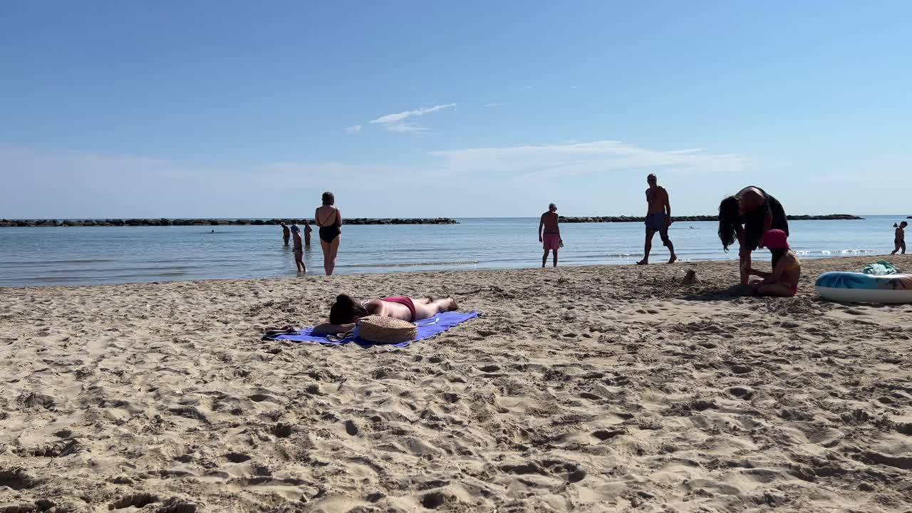 A serene beach scene featuring people relaxing on the sandy shore and swimming in calm waters. Various individuals are sunbathing, playing, and enjoying a sunny day by the seaside